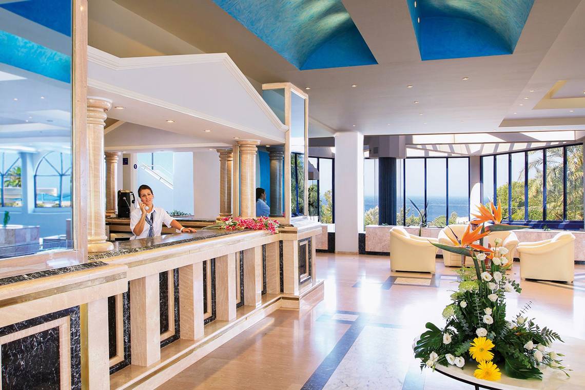 Empfangsbereich des Kresten Palace Hotels mit Rezeptionist, Blumen und Meerblick.