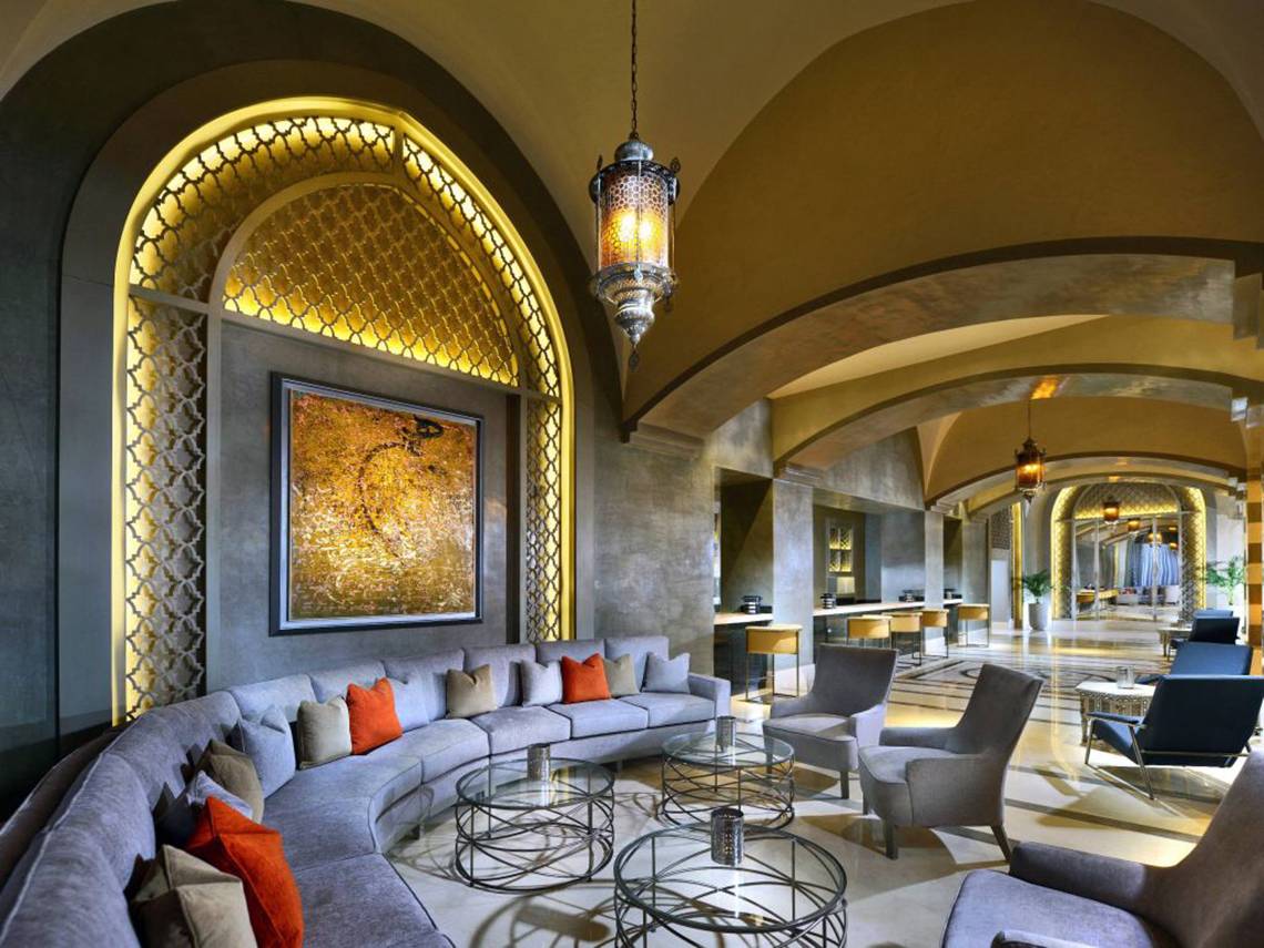 Lobby-Lounge-Bereich des Steigenberger Alcazar Hotels mit einer langen grauen Couch, mehreren Glastischen und einer großen Wandmalerei.