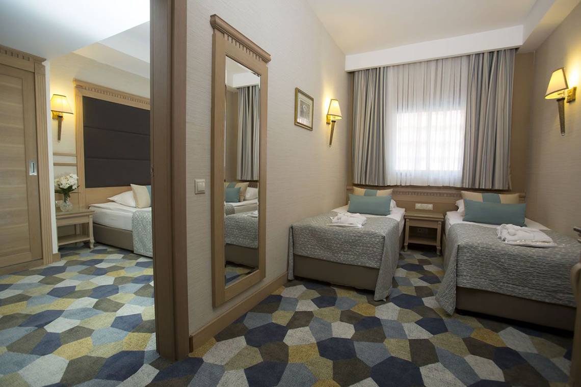 Innenansicht eines Doppelzimmers im Fame Residence Lara & Spa Hotel mit drei Betten, einem Spiegel und einem Tisch mit Blumen.
