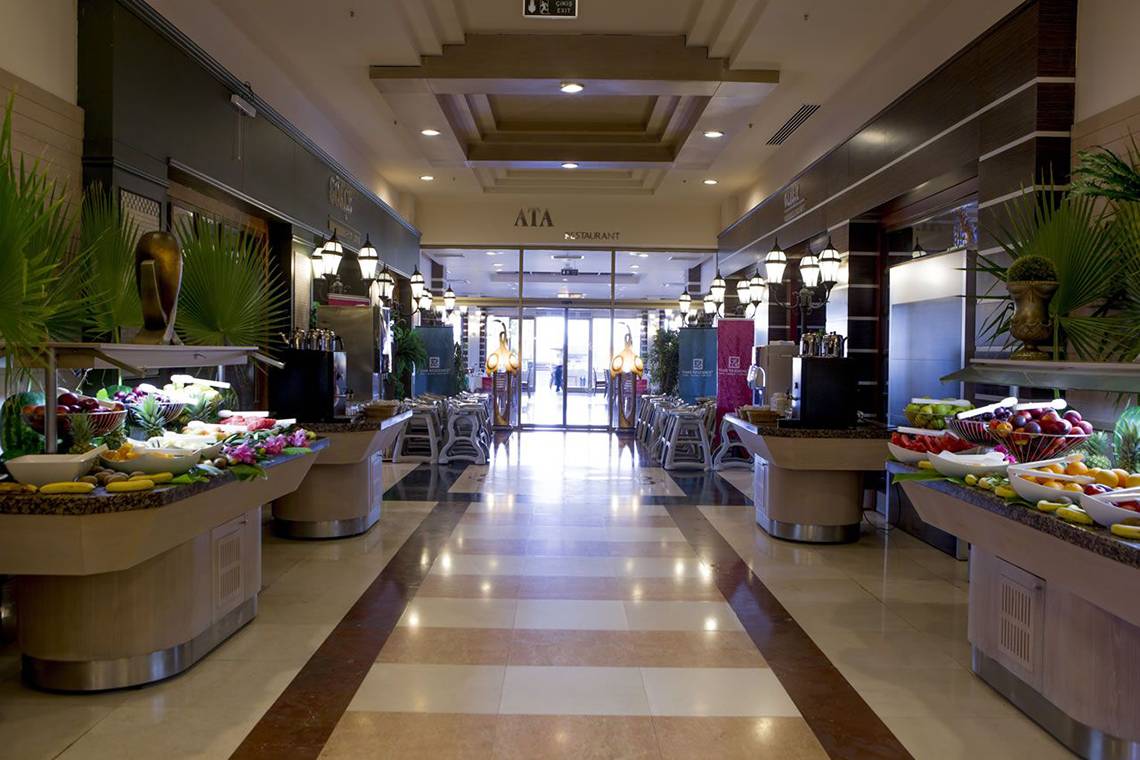 Innenansicht der Halle im Fame Residence Lara & Spa mit Früchtebuffets und beleuchteten Wegen.