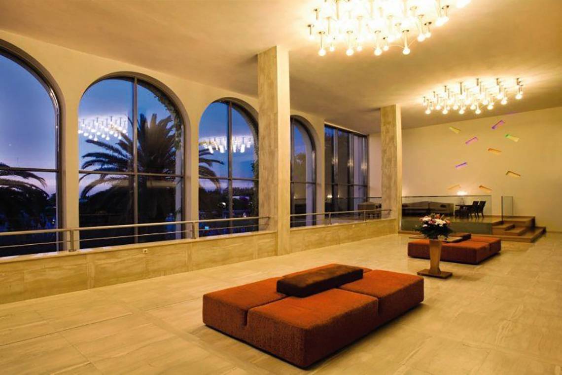 Lobby des Caravia Beach Hotel mit großen Fenstern, Kronleuchtern und Möbeln.