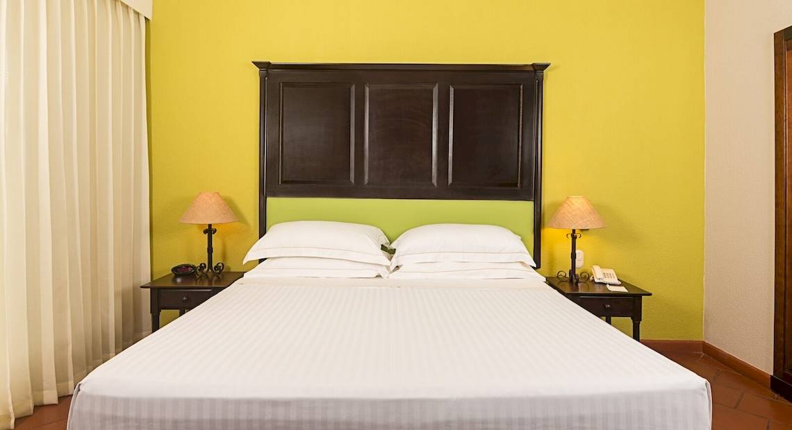 Hotelzimmer im Occidental Papagayo mit gelber Wand, weißem Bett und zwei Nachttischen mit Lampen und Telefonen.