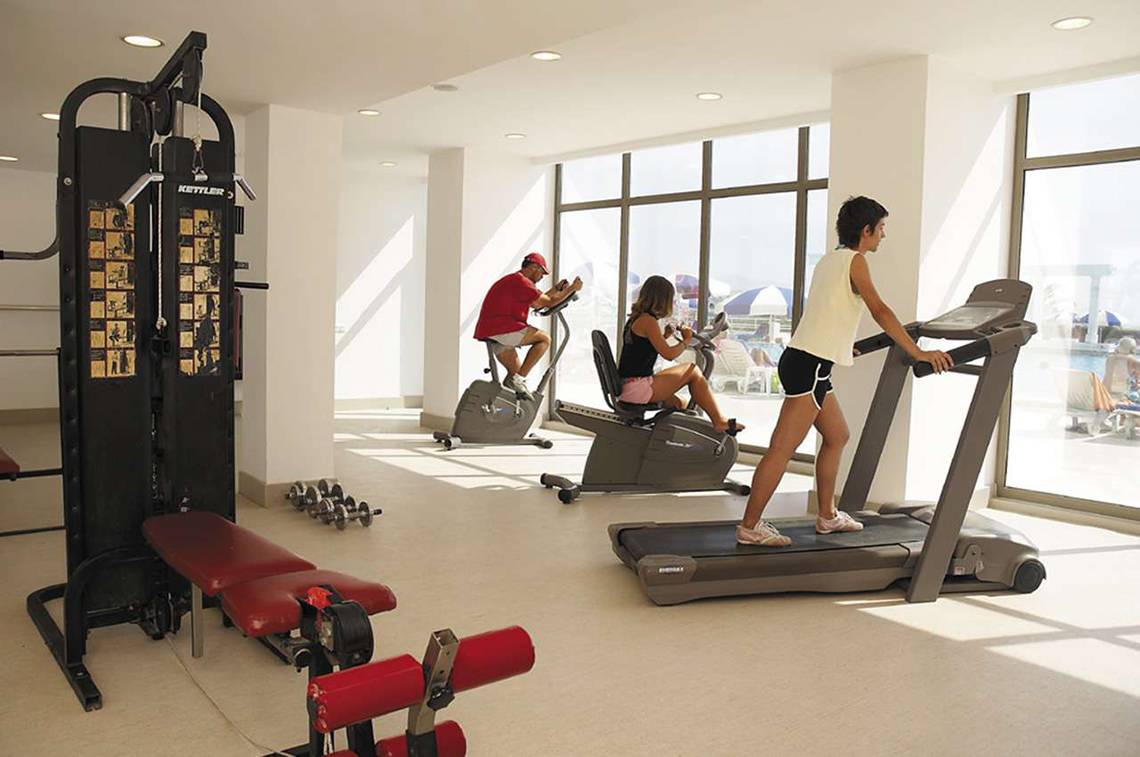 Fitnessstudio im KoruMar Hotel De Luxe mit einer Frau auf einem Laufband und zwei Personen auf anderen Geräten.