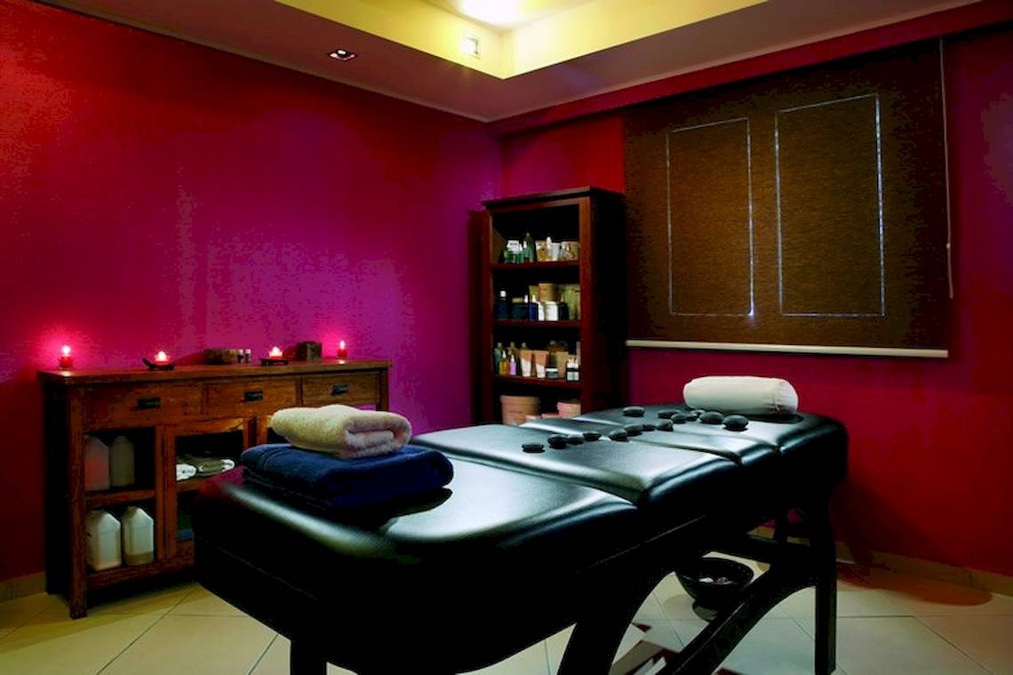 Massageraum im Olympia Golden Beach Resort mit roten Wänden, zwei Massagetischen, Kissen, Kerzen und einem Regal.