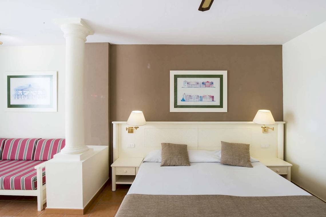Ein schlichtes Hotelzimmer mit einem Bett, einem Sofa, Lampen und zwei Bilderrahmen an der Wand.