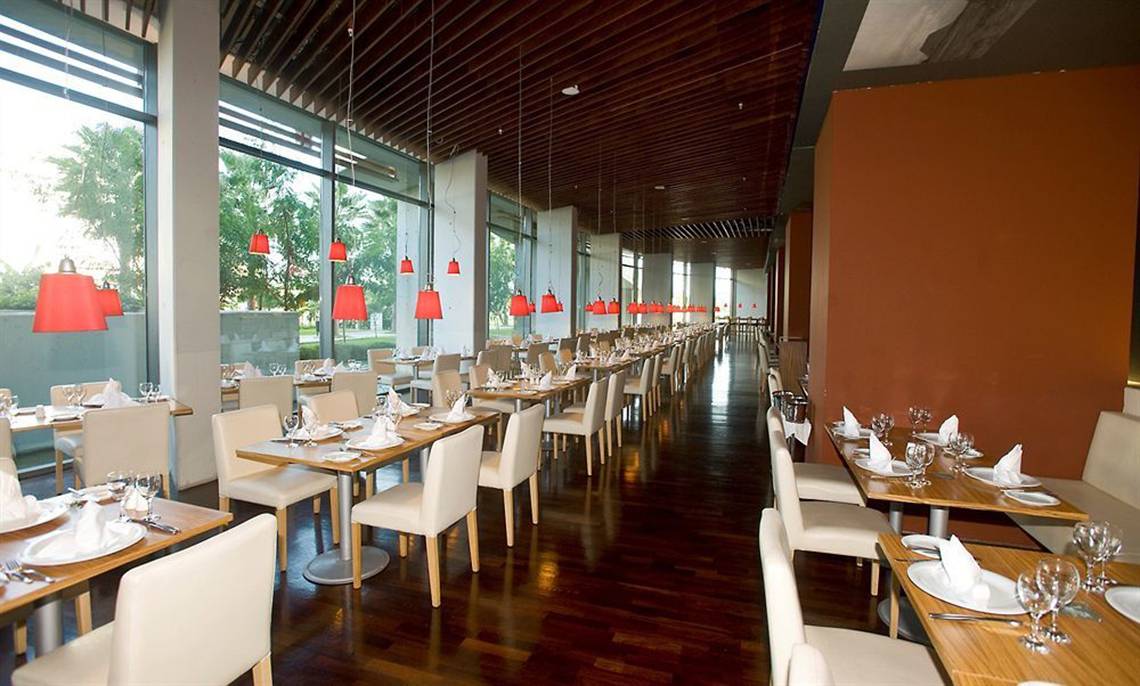 Innenansicht des Restaurants im Nirvana Cosmopolitan Hotel mit langen Reihen von Tischen und Stühlen, roten Lampen und großen Fenstern.