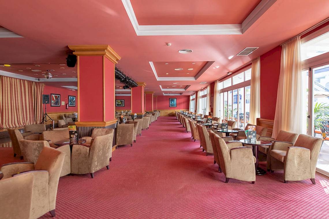 Ein roter Flur im MAC Puerto Marina Hotel mit vielen Sesseln und Tischen, der zu einer Bar führt.