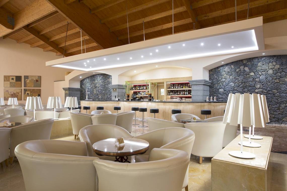 Innenansicht der Lounge-Bar des Hipotels Natura Palace mit weißen Lederstühlen, Holzdecken und Steinwänden.