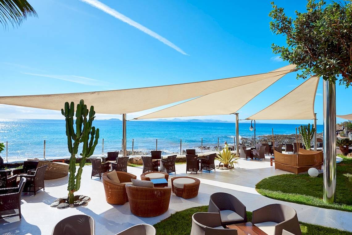 Außenbereich des Hipotels Natura Palace mit mehreren Rattanstühlen und Tischen, überdacht von Sonnenschirmen, mit Blick auf das Meer.