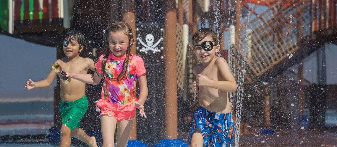 Drei Kinder laufen durch Wasserspritzer im Wasserpark des DoubleTree by Hilton Resort & Spa Marjan Island.
