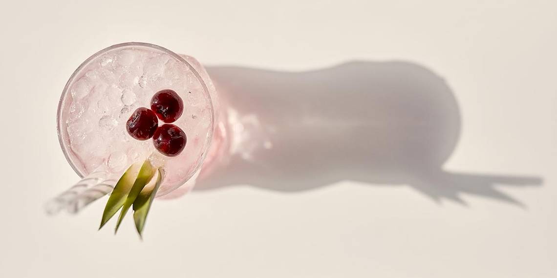 Cocktail mit Eis, roten Beeren und grünem Gras im Glas mit Schatten auf weißer Oberfläche