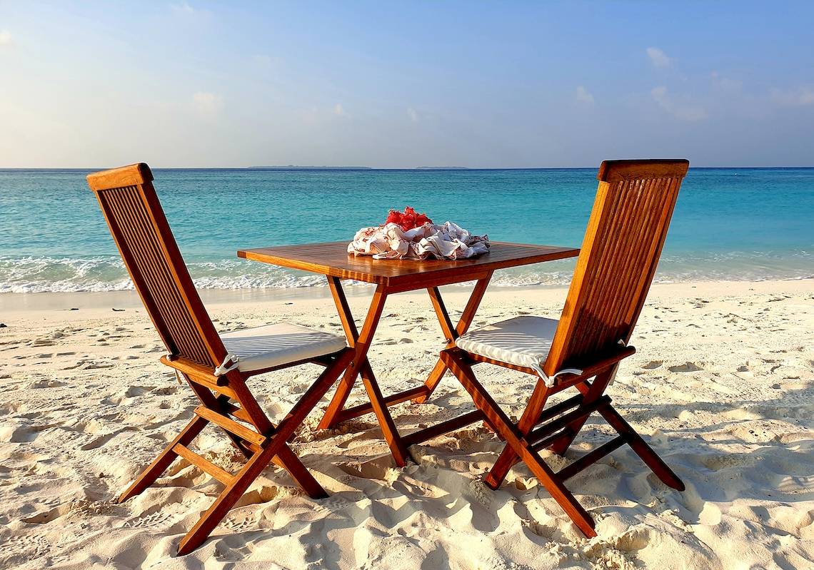 Ein Holztisch am Strand des Barefoot Eco Hotels mit zwei Stühlen und Blick auf das Meer.
