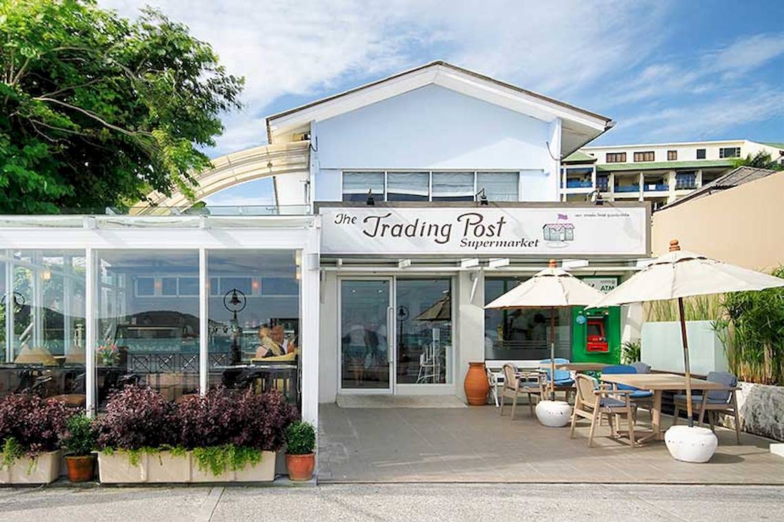Außenansicht des The Trading Post Supermarkts mit Sitzplätzen und Sonnenschirmen.