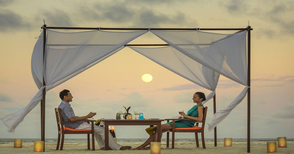 Ein Paar sitzt unter einem Baldachin am Strand und genießt ein romantisches Abendessen bei Sonnenuntergang im Angsana Velavaru.