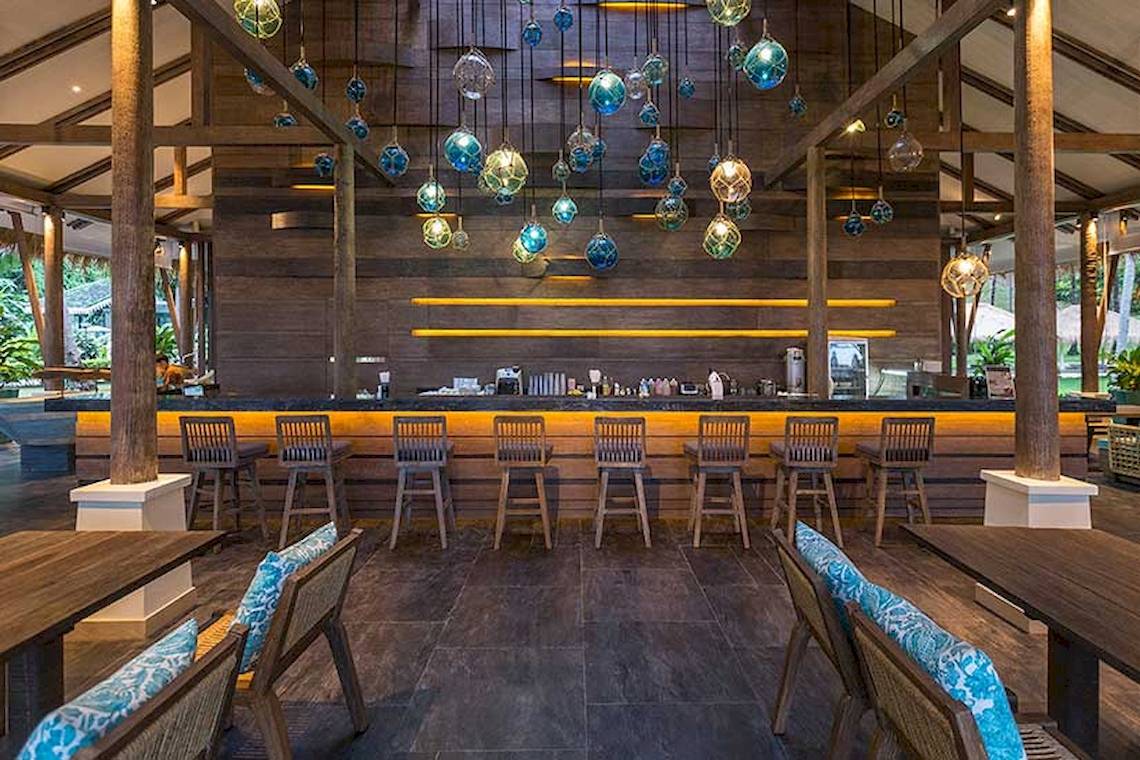 Innenansicht der Bar im Cape Panwa Hotel mit Holzmöbeln und blauen Glasleuchten.