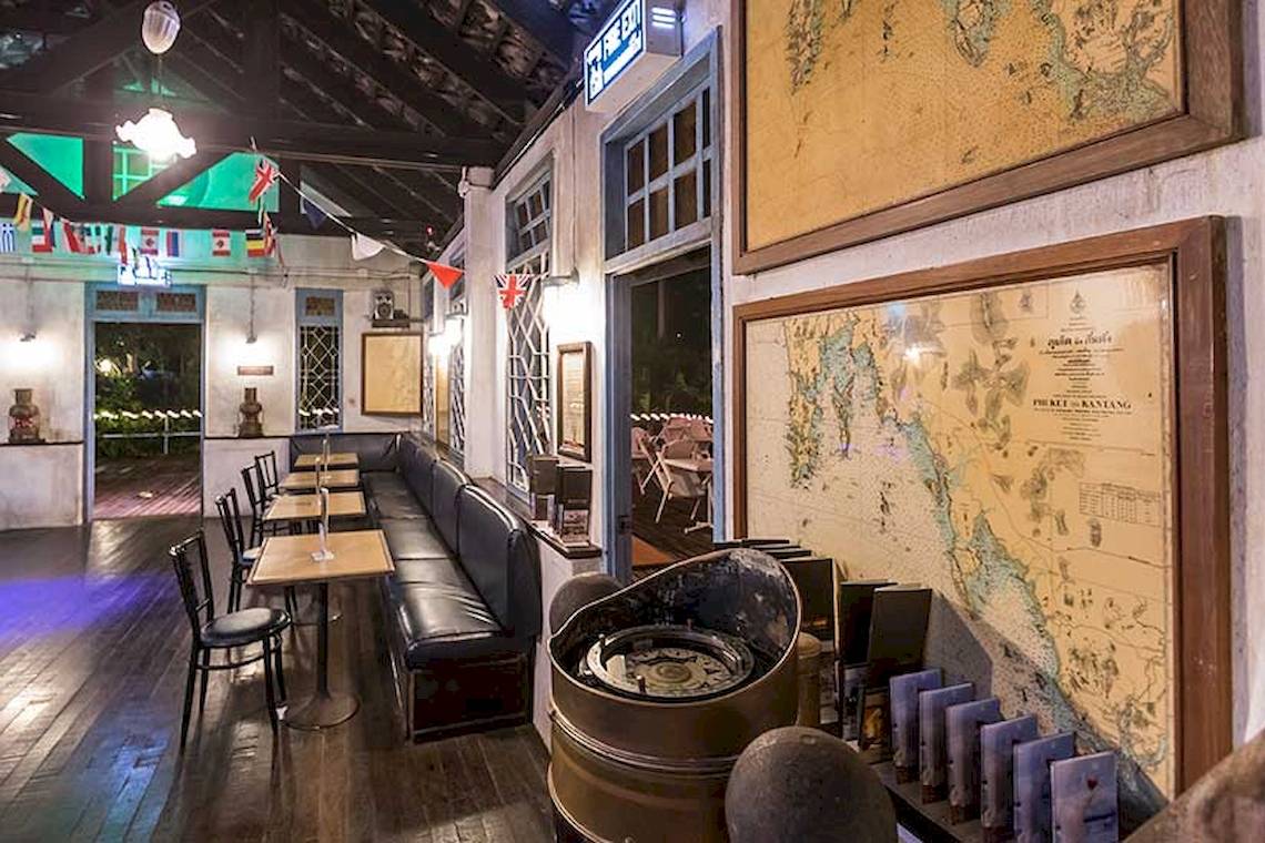 Innenansicht des Cape Panwa Hotels mit einem langen schwarzen Ledersofa, Tischen und Stühlen, alten Karten und maritimen Dekor.