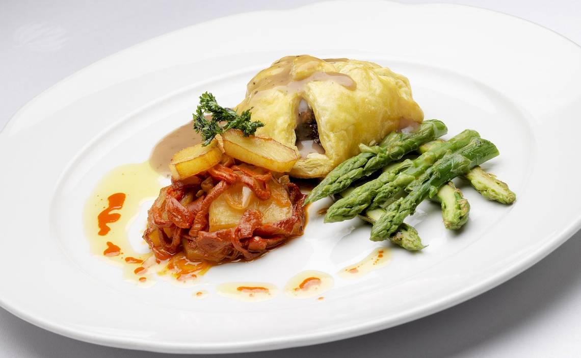 Ein Teller mit einem Gericht, bestehend aus einem gebackenen Teigtaschen, gebratenen Kartoffeln, Spargel und Sauce.