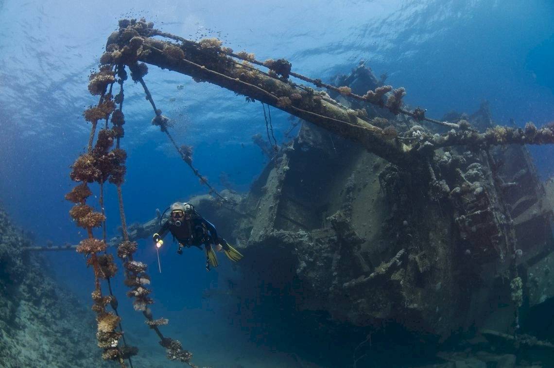 Taucher in Tauchausrüstung bei einem verrosteten Schiffswrack unter Wasser.