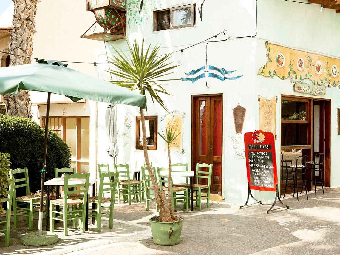 Terrasse des Blue Sea Beach Hotels mit grünen Stühlen, Tischen und einem Palmenbaum.