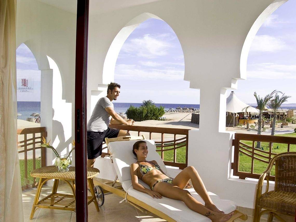 Eine Frau im Bikini liegt auf einer Liege auf dem Balkon mit Blick auf das Meer und einen Mann der sie beobachtet.