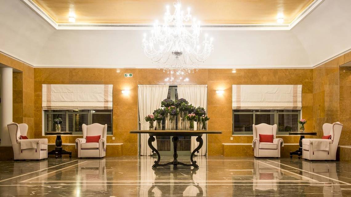 Empfangsbereich des Bella Beach Hotels mit einem großen Kronleuchter, weißen Sesseln und einem Tisch mit Blumen.