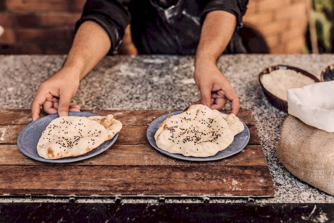 Ein Koch präsentiert zwei gebackene Fladenbrote mit Sesam auf einem Holzbrett.