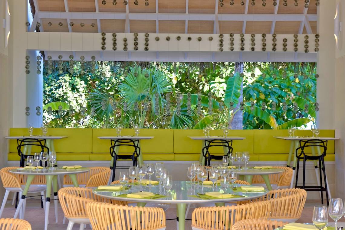 Innenbereich des Kandima Maldives Hotels mit gelben Möbeln und einer großen Fensterfront, die auf einen tropischen Garten hinausblickt.