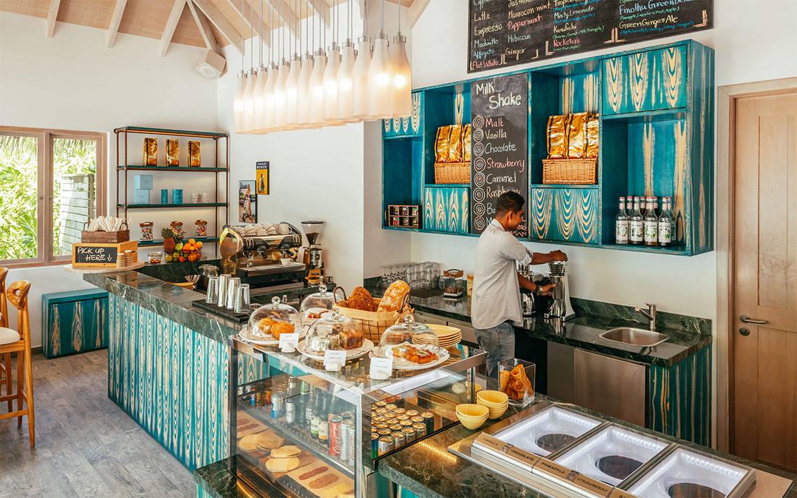 Blick in das Café im Finolhu Baa Atoll Maldives mit einem Mann, der Getränke zubereitet und einer Theke mit vielen verschiedenen Broten und Desserts.