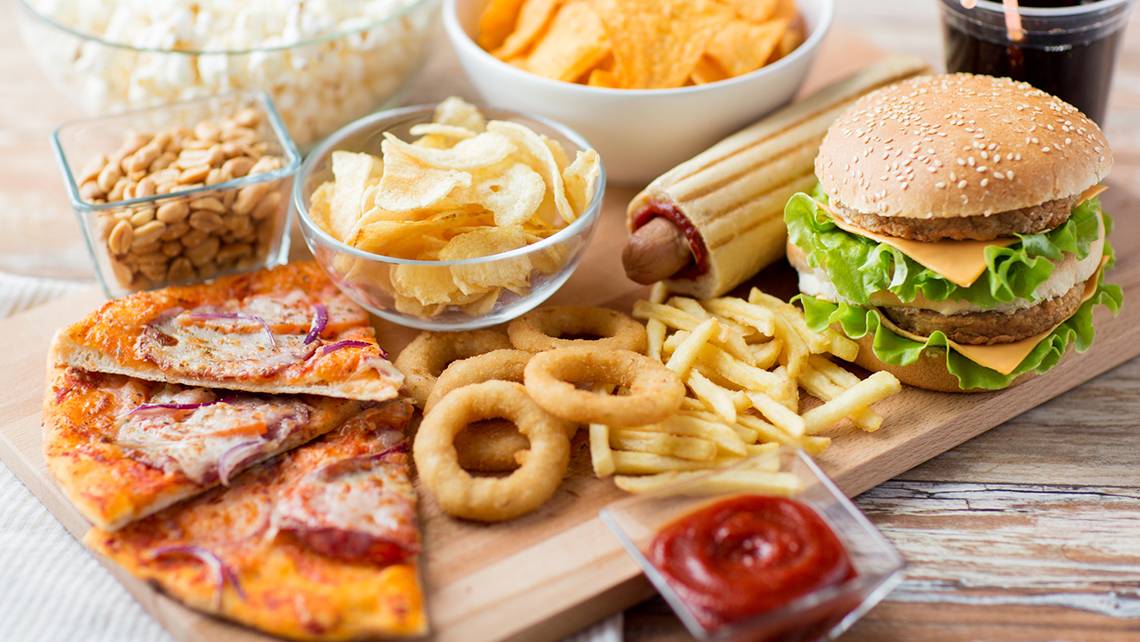 Eine Auswahl an ungesundem Essen wie Pommes, Pizza, Burger, Chips und Soda auf einem Holztisch.