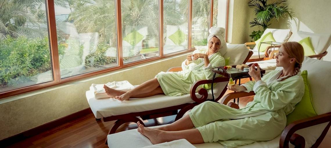 Zwei Frauen im grünen Bademantel sitzen in einem Hotel-Loungebereich mit Blick auf den See.