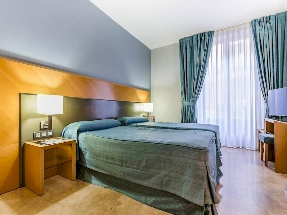 Doppelbettzimmer im Del Mar - Barcelona Hotel mit blauen Vorhängen und Holzmöbeln.