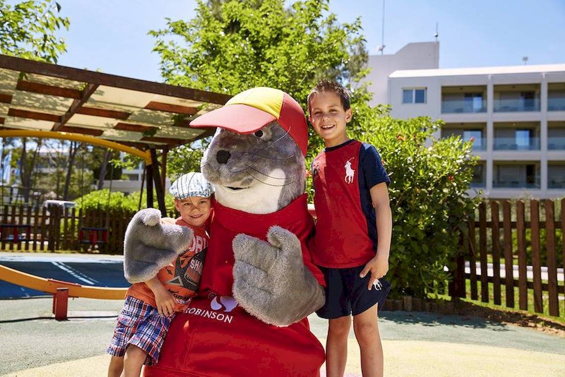 Zwei Kinder stehen neben dem Maskottchen von Robinson Kyllini Beach Hotel im Spielplatz.