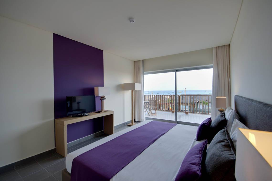 Innenansicht eines Zimmers im Oasis Salinas Sea Hotel mit violetter Wand, Fernseher und Balkon mit Meerblick.