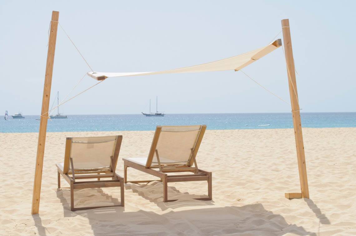Zwei Holzliegen unter einem Sonnensegel am Strand mit Meer im Hintergrund