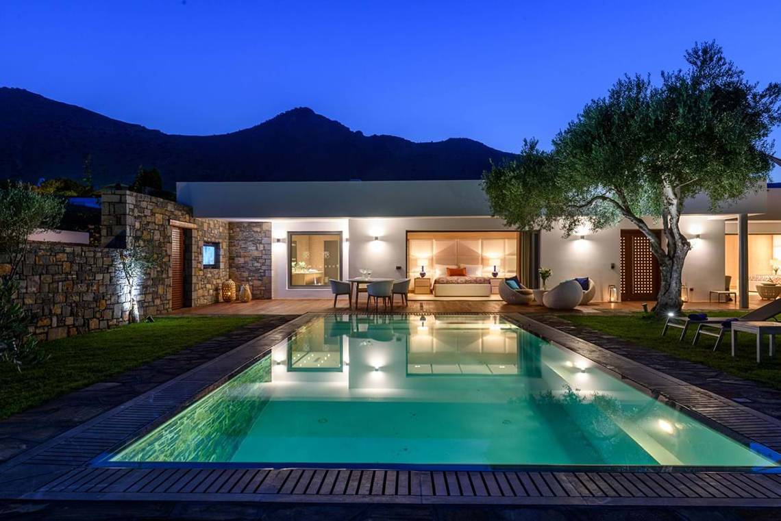 Außenbereich des Elounda Beach Hotel & Villas bei Nacht, mit beleuchtetem Swimmingpool und Blick auf Berge.
