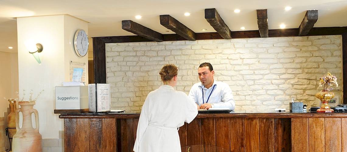 Ein Mitarbeiter der Rezeption des Djerba Resorts hilft einer Frau in einem weißen Bademantel an der Rezeption.