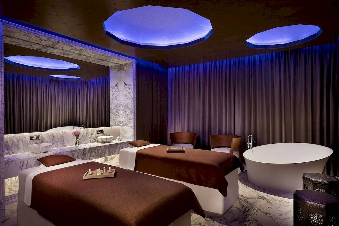 Spa-Behandlungsraum im W Muscat Hotel mit zwei Massagetischen, einem Badewanne, blauen Deckenleuchten und Vorhängen.