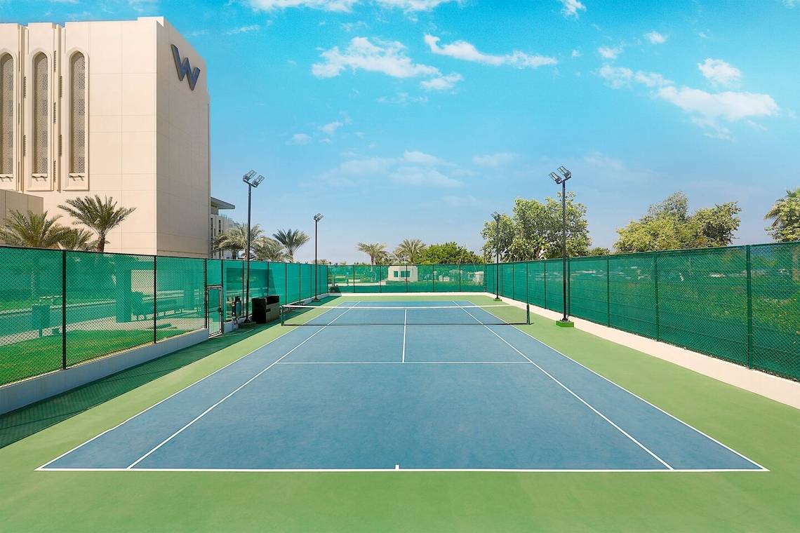 Tennisplatz vor dem W Muscat Hotel mit grünen Zäunen und Gebäude im Hintergrund.
