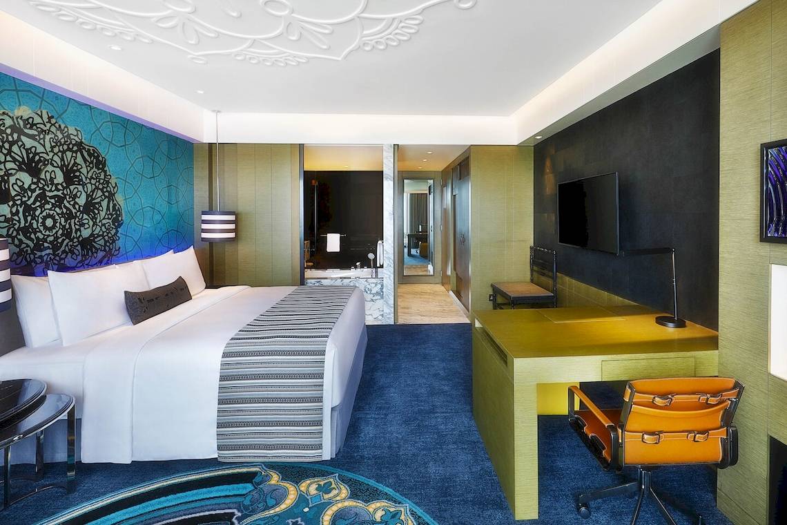 Innenansicht der Suite im W Muscat Hotel mit einem großen Bett, Fernseher und Arbeitsbereich.