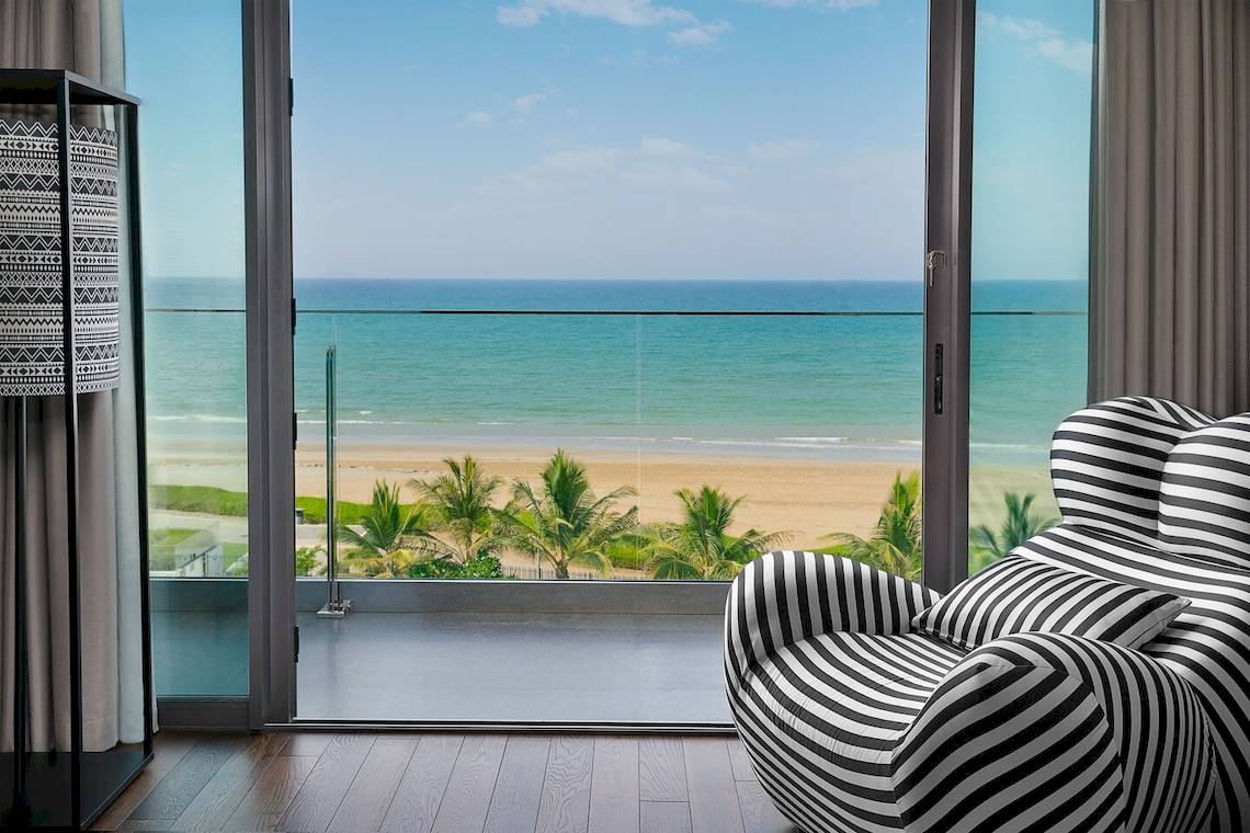Blick aus einem Hotelzimmer auf das Meer mit einer schwarzen und weißen gestreiften Liege und einem Glasfenster.