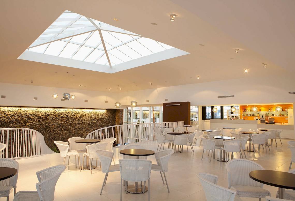 Innenbereich des Be Live Experience Lanzarote Beach Hotels mit vielen Tischen und Stühlen, hoher Glaskuppel und Steinwand.