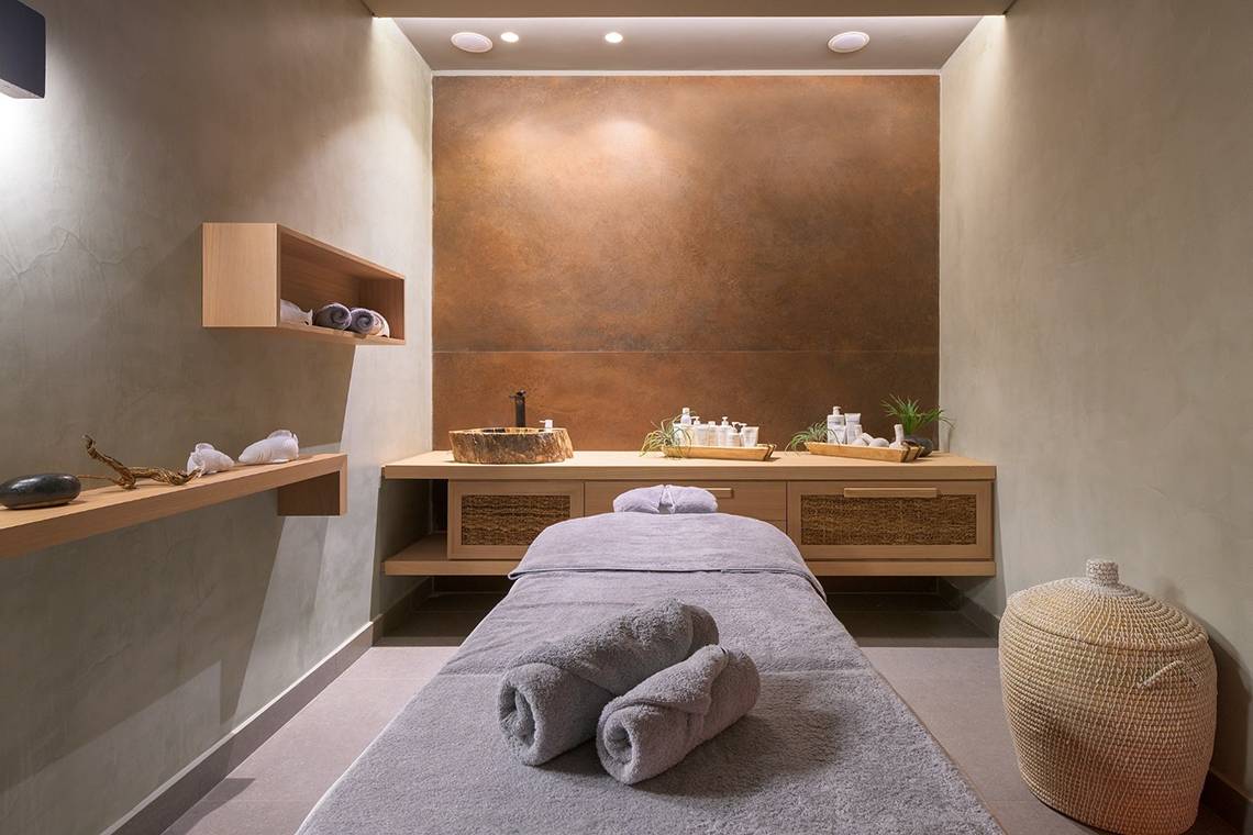 Massagebereich im Olympic Palace Resort mit einem Massagetisch, zwei Handtüchern, einem Holzregal, einem Waschbecken und einer Kupferwand.