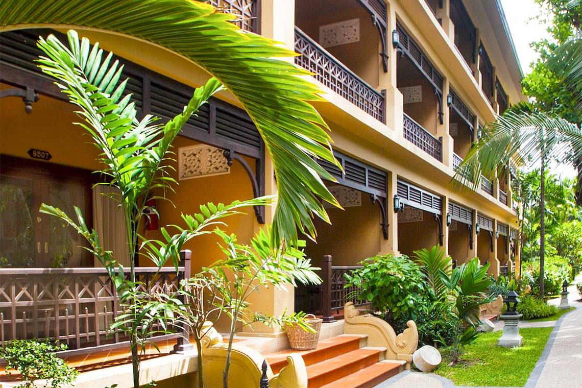 Außenansicht des Fair House Beach Resorts mit gepflegtem Garten, Palmen und gelber Fassade.