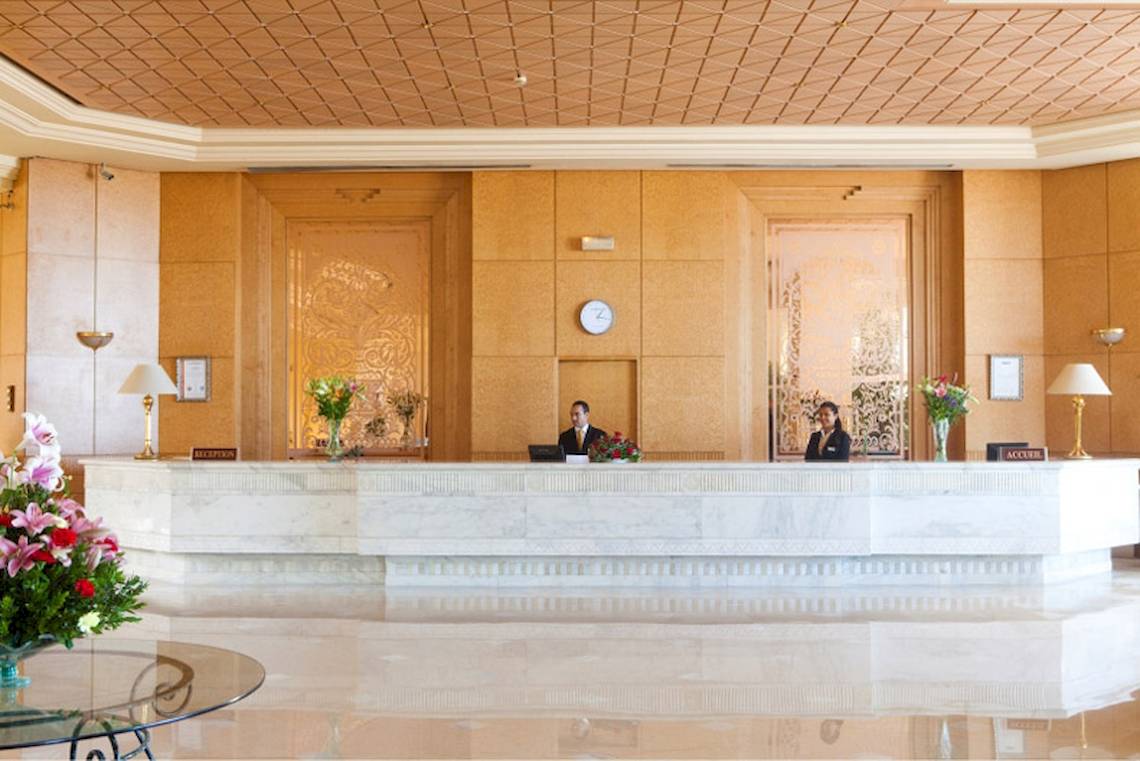 Rezeptionsbereich des El Mouradi Hammamet Hotels mit zwei Mitarbeitern und Blumenarrangements.