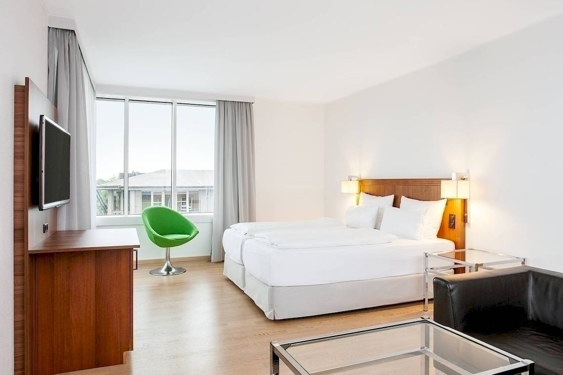 Innenansicht eines Hotelzimmers im NH Hamburg Altona mit zwei Betten, Fernseher, Schreibtisch und grünem Stuhl.