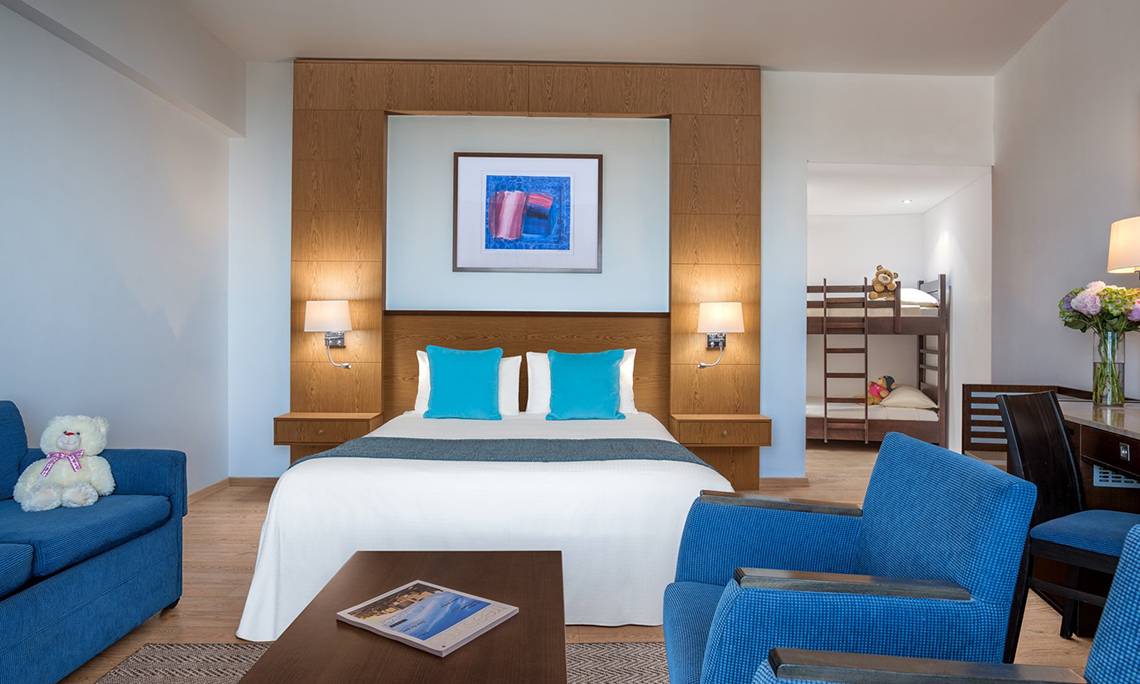 Innenansicht eines Zimmers im Mediterranean Beach Hotel in Zypern mit einem Bett, zwei blauen Kissen, einem Sofa, einem Tisch, einem Teddybär und einem Bild an der Wand.