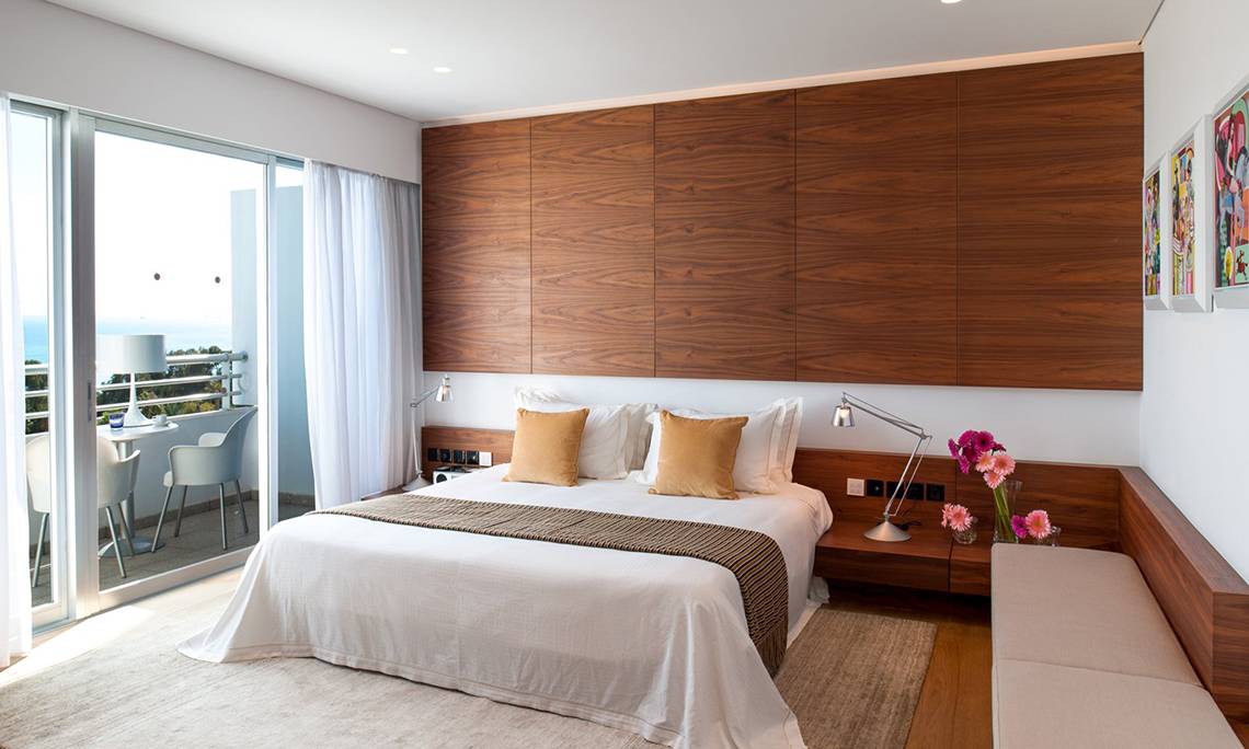 Schlafzimmer mit Balkon im Mediterranean Beach Hotel in Zypern, mit modernen Holzwänden, einem großen Bett und einem Ausblick auf das Meer.