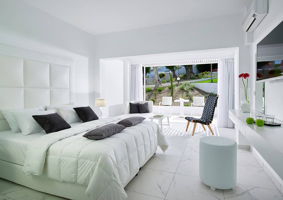 Schlafzimmer im Dimitra Beach Hotel & Suites mit weißem Bett und Möbeln, Balkonzugang und Gartenblick.