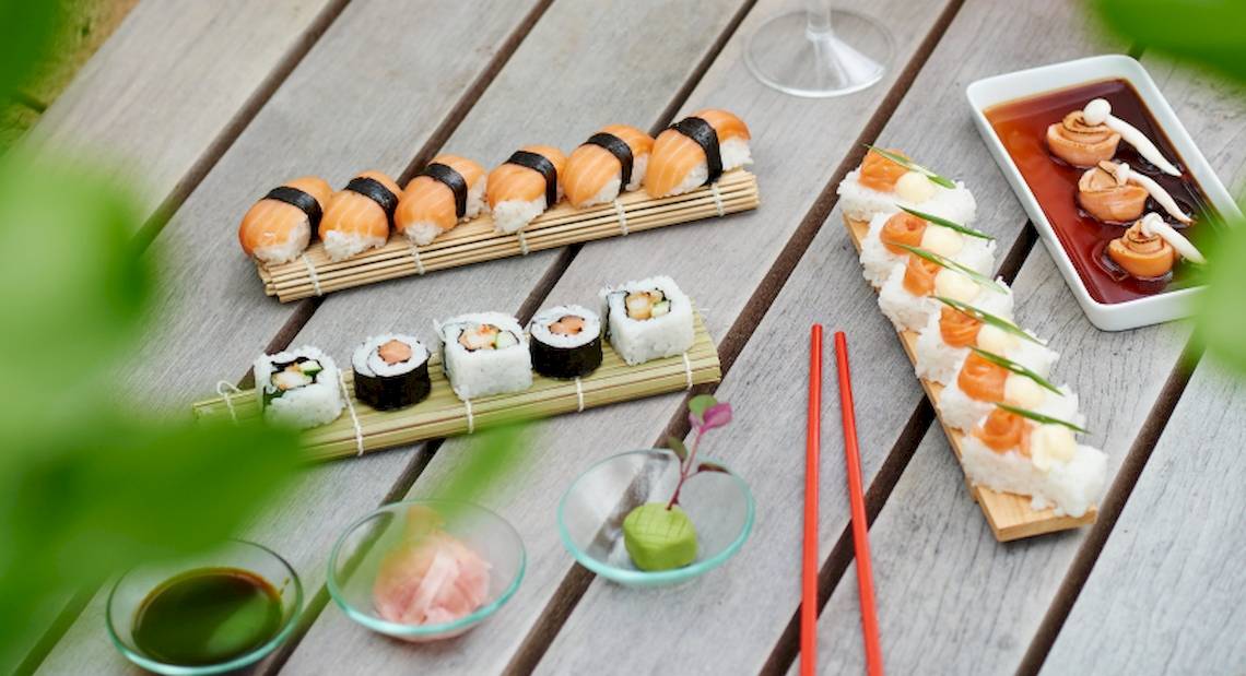 Mehrere Sushi-Platten und Beilagen auf einem hölzernen Tisch, darunter ein Glas Wein und verschiedene Saucen.