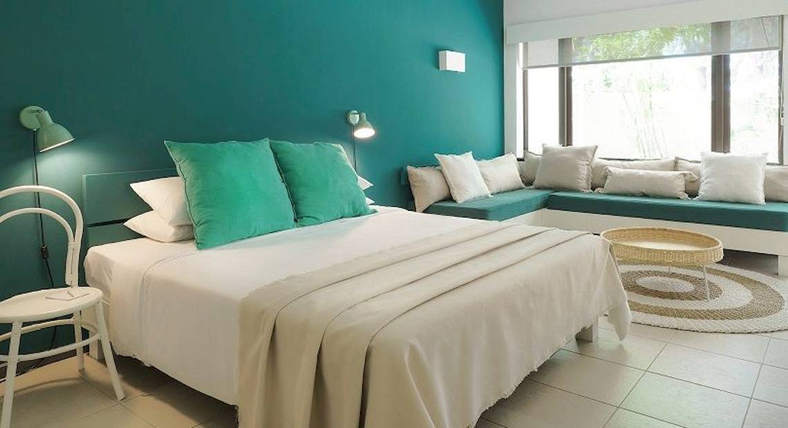 Ein türkisfarbenes Zimmer mit einem großen Bett, grünen Kissen und weißen Decken, einem weißen Stuhl und einer Couch in Blau. Neben dem Bett befindet sich ein Tisch mit einer Tischlampe.
